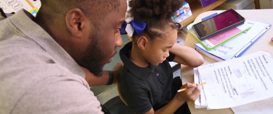 Video about UB Teacher Residency Program; photo of teacher helping student with schoolwork at desk. 